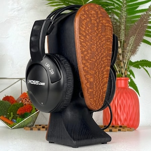 Op de afbeelding: Zwarte Koss UR.20 over-ear hoofdtelefoon rustend op een gestileerde houten hoofdstandaard. De standaard heeft een donkerzwarte basis en een getextureerde, lichtbruine houtnerf voorplaat. De hoofdtelefoon heeft een zwart kunststof frame.