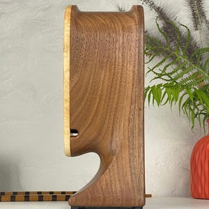 May include: A side view of a wooden clock featuring a dark brown walnut body with a lighter, pale yellow wood face. The clock has a curved, sculptural silhouette with a cutout base and a visible horizontal slot on the side of the light wood face.