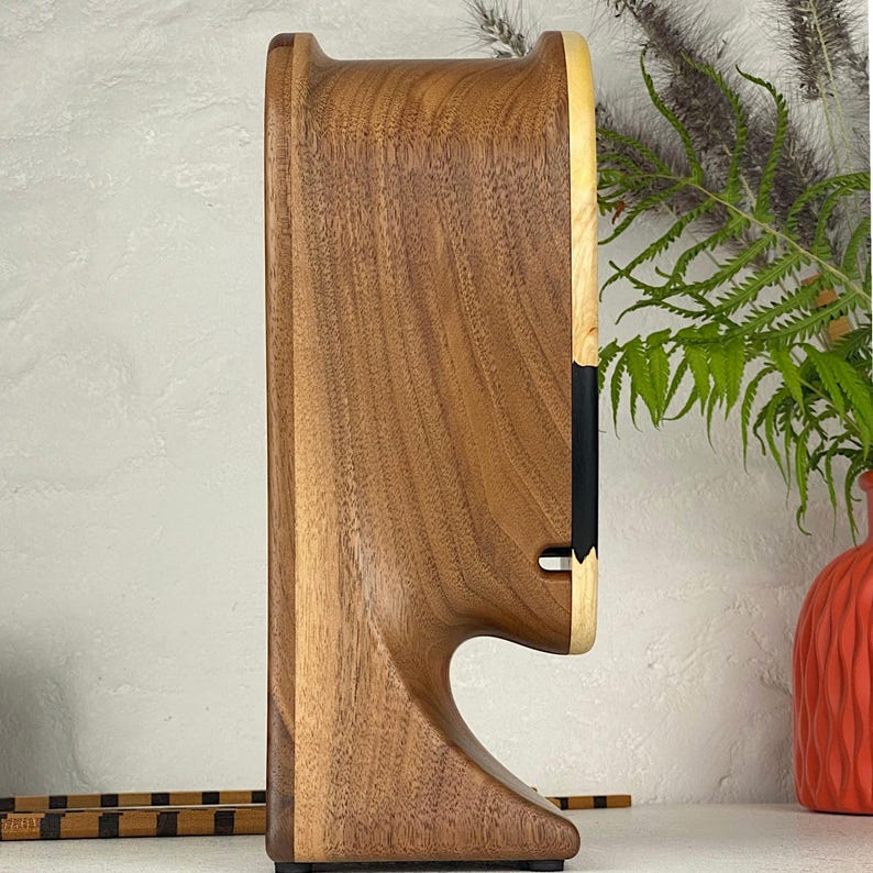 May include: A vertical wooden speaker with a curved, sculpted profile made from dark brown walnut wood. The front face features a lighter-toned wood border with a black rectangular inlay near the centre and a small horizontal cutout slot below it.