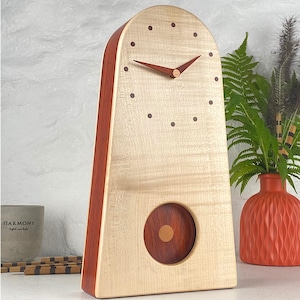 Op de afbeelding: Een hoge, boogvormige houten klok met een lichtgekleurde, generfde wijzerplaat en donkere, ronde uurmarkeringen. De klok heeft twee donkere, puntige wijzers en een ronde opening onderaan die een donkerhouten slinger met een schijfje toont.