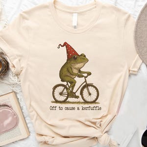 May include: Cream-colored t-shirt featuring a whimsical illustration of a frog riding a bicycle, wearing a red and white polka-dotted hat. The text "Off to cause a kerfuffle" is printed below the image. The shirt is knotted at the bottom.