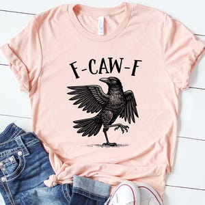 May include: A light pink t-shirt with the text "F-CAW-F" above a black crow illustration. The shirt is paired with blue jeans and white sneakers.