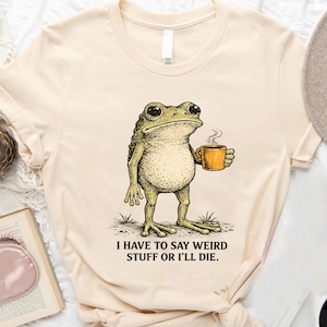 May include: Cream-colored t-shirt featuring a detailed illustration of a frog holding a steaming orange mug. The frog stands on grass, and the text "I HAVE TO SAY WEIRD STUFF OR I'LL DIE" is printed below the frog.