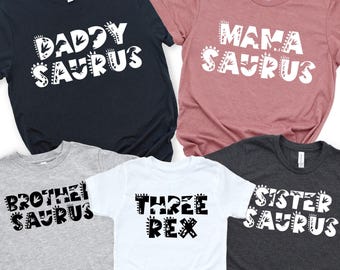 May include: Five short-sleeved t-shirts with dinosaur-themed text. The shirts are navy, mauve, gray, white, and charcoal. Text includes "Daddy Saurus", "Mama Saurus", "Brother Saurus", "Three Rex", and "Sister Saurus". A toy dinosaur is in the bottom right.
