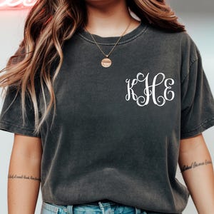 May include: Dark gray t-shirt with a white monogram design. The shirt has a classic crew neck and short sleeves. The monogram features the letters K, H, and E in a decorative script font. The shirt is made of a soft, comfortable material.