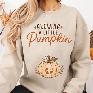 May include: A sand-colored crewneck sweatshirt featuring the text "GROWING A LITTLE Pumpkin" in brown lettering. Below the text is a graphic of a light orange pumpkin with two small baby footprints centered on it, surrounded by small leaf accents.