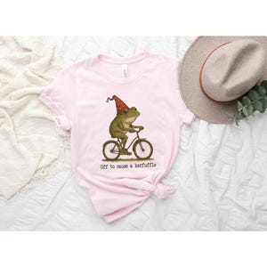 Op de afbeelding: Lichtroze t-shirt met een grillige illustratie van een kikker op een fiets, met een rode feesthoed. De tekst "Off to cause a kerfuffle" staat eronder. Op de achtergrond is een beige hoed te zien.