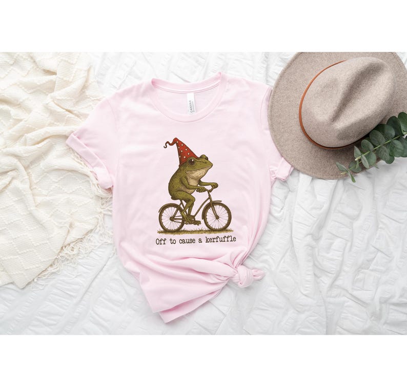 Op de afbeelding: Lichtroze t-shirt met een grillige illustratie van een kikker op een fiets, met een rode feesthoed. De tekst "Off to cause a kerfuffle" staat eronder. Op de achtergrond is een beige hoed te zien.