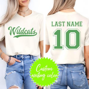 Puede incluir: Camiseta color crema con la palabra "Wildcats" en verde en la parte delantera. La parte trasera muestra "LAST NAME" sobre el n&uacute;mero "10" en verde. La imagen tambi&eacute;n incluye un gr&aacute;fico verde ne&oacute;n que dice "Custom writing color".