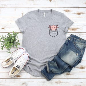 May include: A heather gray t-shirt featuring a pocket design with a cartoon axolotl. The axolotl is pink with a smiling face. The shirt is paired with blue jeans and white sneakers with red accents.