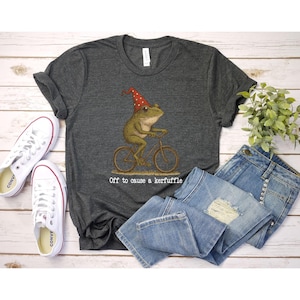 Op de afbeelding: Donkergrijs t-shirt met een kikker op een fiets met een rode hoed. De tekst "Off to cause a kerfuffle" staat eronder. Het shirt wordt gecombineerd met een blauwe jeans en witte canvas sneakers.
