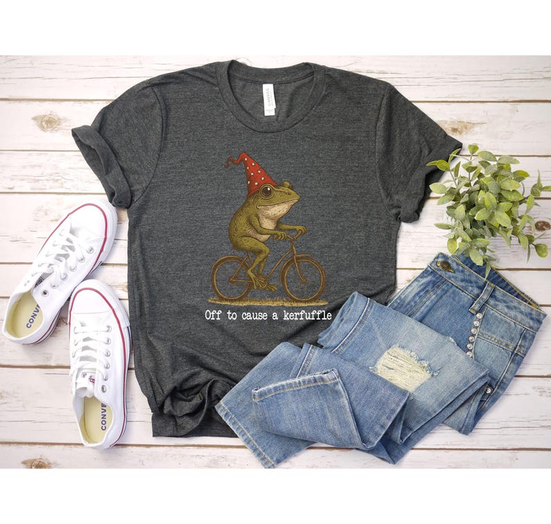 Op de afbeelding: Donkergrijs t-shirt met een kikker op een fiets met een rode hoed. De tekst "Off to cause a kerfuffle" staat eronder. Het shirt wordt gecombineerd met een blauwe jeans en witte canvas sneakers.