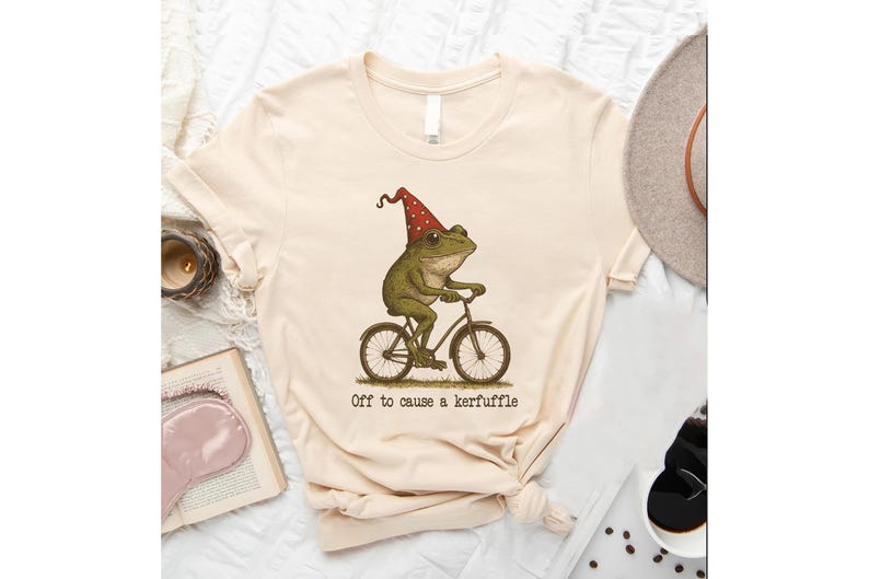 Op de afbeelding: Cr&egrave;mekleurig T-shirt met een illustratie van een kikker op een fiets, met een rood-witte stippenhoed. De tekst "Off to cause a kerfuffle" staat eronder. Het T-shirt is aan de onderkant geknoopt.