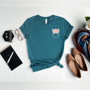May include: Teal t-shirt with a pocket design featuring a pink axolotl. The shirt is styled with a black notebook, glasses, jewelry, and a pair of brown shoes. The shirt is made of a soft, comfortable material.