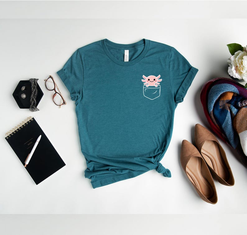 May include: Teal t-shirt with a pocket design featuring a pink axolotl. The shirt is styled with a black notebook, glasses, jewelry, and a pair of brown shoes. The shirt is made of a soft, comfortable material.