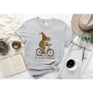 Op de afbeelding: Lichtgrijs t-shirt met een illustratie van een kikker op een fiets met een rode en gouden feesthoed. De tekst "Off to cause a kerfuffle" staat eronder. Het t-shirt is opgevouwen en ligt op een witte ondergrond.