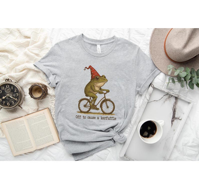 Op de afbeelding: Lichtgrijs t-shirt met een illustratie van een kikker op een fiets met een rode en gouden feesthoed. De tekst "Off to cause a kerfuffle" staat eronder. Het t-shirt is opgevouwen en ligt op een witte ondergrond.