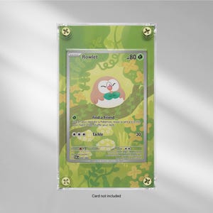 May include: A framed Pokémon trading card featuring Rowlet, an owl-like creature, perched on a branch. The card is set against a green and yellow floral background, secured by four gold-colored screws. The text "Card not included" is at the bottom.