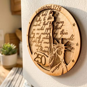 Puede incluir: Un imán redondo de madera con una imagen grabada de la Estatua de la Libertad. El imán incluye el texto "Established 1776" y una cita de la Declaración de Independencia. También es visible el número "250".