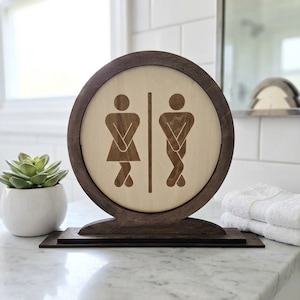 May include: A wooden restroom sign with a circular design. The sign features a light-colored center with etched figures representing male and female restrooms. The frame and base are dark brown. A small succulent in a white pot is to the left.