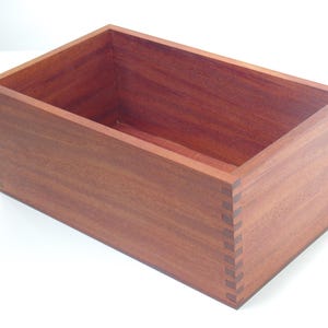 May include: A rectangular wooden box with a natural reddish-brown finish. The box is made of solid wood and has a simple, elegant design.