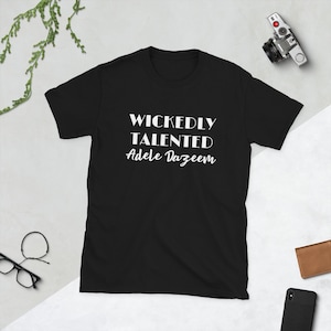 以下が含まれることがあります： 白文字で「WICKEDLY TALENTED Adele Dazeem」と書かれた黒いTシャツ。半袖Tシャツはクルーネックです。テキストは太字のサンセリフ体です。カジュアルな普段着です。