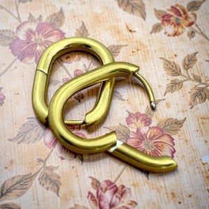 May include: A pair of gold-toned hoop earrings with an elongated, rounded rectangular shape. The earrings feature a smooth, polished metallic finish and a hinged post mechanism that secures the hoop closed, creating a continuous, sleek loop.