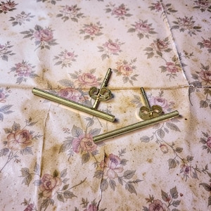 May include: A pair of gold-colored bar stud earrings. The earrings are displayed on a floral patterned paper with pink and purple roses. The earrings have a simple, minimalist design.