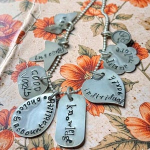 Peut inclure: Un collier à breloques en argent avec divers pendentifs. Les pendentifs comprennent un cœur, une étiquette, une étoile et une fleur, chacun gravé de mots tels que "intégrité" et "connaissance". Le collier est présenté sur un fond floral.