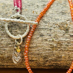 May include: A handcrafted necklace featuring a silver toggle clasp and a pendant with heart details. The necklace has two strands of pink beads and a strand of orange beads. The jewelry is displayed on a textured wooden surface.