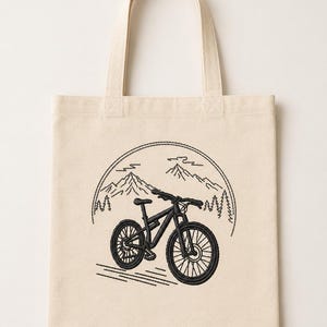 Peut inclure: Un sac fourre-tout en toile beige avec une broderie noire représentant un vélo tout-terrain. Le motif comprend un contour circulaire encadrant une chaîne de montagnes avec des sapins en arrière-plan et des lignes horizontales sous le vélo.