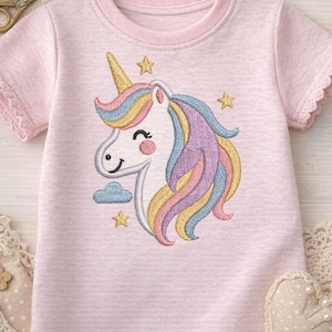 May include: A light pink short-sleeved t-shirt featuring an embroidered unicorn design. The unicorn has a golden horn, a white face, and a colorful mane in shades of yellow, blue, and purple. The design includes stars and a cloud.