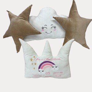 May include: A collection of baby pillows in the shape of a cloud, stars, and a crown. The cloud and crown pillows have embroidered details, including a rainbow and the name "Amira Ana-Maria". The star pillows are a solid brown color.