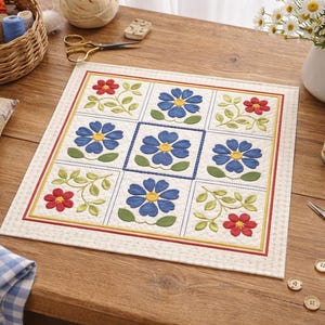 Op de afbeelding: Een vierkant, gewatteerd bloemenontwerp met blauwe en rode bloemen, groene bladeren en gele accenten. Het ontwerp is omgeven door een rode en gele rand. Naaibenodigdheden zoals garen, schaar en knopen zijn ook aanwezig.