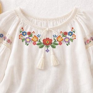 May include: White blouse with embroidered floral design in red, orange, blue, and purple. The blouse features a round neckline with lace trim, long sleeves, and decorative tassels. A straw hat is visible in the upper left corner.