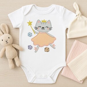 May include: White baby bodysuit featuring a grey cat wearing a crown and a peach dress, holding a gold star wand. The bodysuit has a snap closure and is decorated with embroidered geometric shapes and a small box. A cream hat and pink and cream blankets are also in the image.