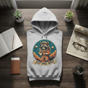 May include: A light gray hoodie featuring a cartoon raccoon in an astronaut suit, holding a cup. The design is set in a blue circle with the words "DEEP SPACE DIVER!" on a banner. The hoodie is on a wooden surface.