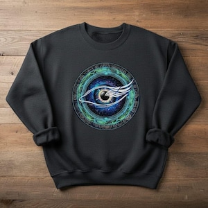 May include: A black crewneck sweatshirt featuring a detailed eye design. The eye is set within a circular frame with intricate patterns and a celestial background. The eye has a wing design on the side.