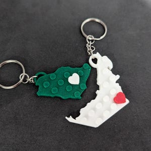 Può includere: Due portachiavi con ciondoli a forma di mappa dalla superficie testurizzata a blocchi. Un ciondolo è verde scuro con un piccolo cuore bianco, l'altro è bianco con un piccolo cuore rosso. Entrambi includono anelli portachiavi in metallo argentato.