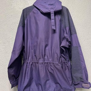 May include: A purple and gray hooded jacket with a drawstring waist and adjustable cuffs. The jacket has a full zipper and a small pocket on the sleeve. The back of the jacket is visible, hanging from a metal hook.