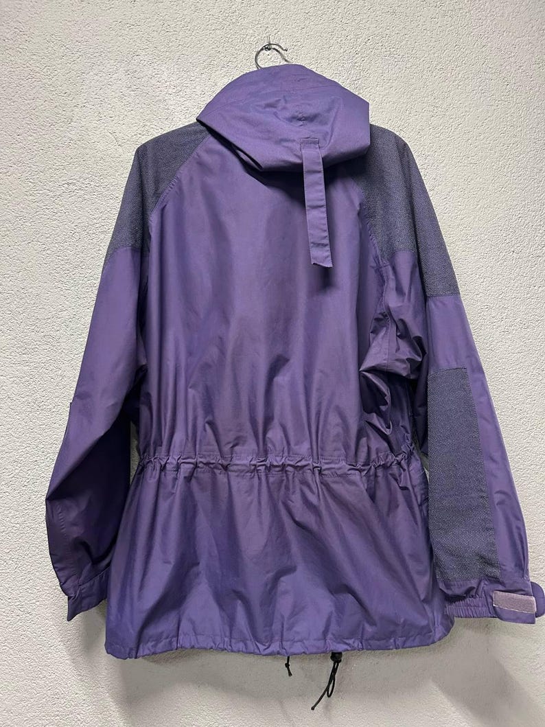May include: A purple and gray hooded jacket with a drawstring waist and adjustable cuffs. The jacket has a full zipper and a small pocket on the sleeve. The back of the jacket is visible, hanging from a metal hook.