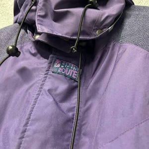 May include: Close-up of a purple jacket with a hood. The jacket has a drawstring and a small logo that reads "BAHIA ROUTE". The jacket is made of a smooth, water-resistant material.