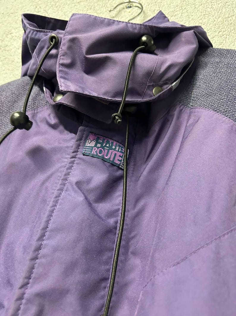 May include: Close-up of a purple jacket with a hood. The jacket has a drawstring and a small logo that reads "BAHIA ROUTE". The jacket is made of a smooth, water-resistant material.