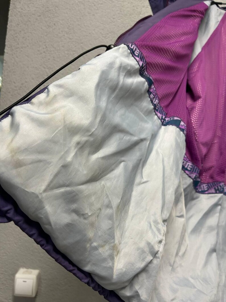 May include: Interior view of a purple and white jacket. The jacket's lining is white, with a purple mesh panel visible. The jacket has a drawstring and a printed border with the word "HATE" repeated. The jacket appears to be worn.