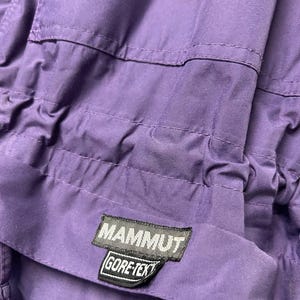 May include: Close-up of a purple garment with stitched seams and a drawstring. A black and white label reads "MAMMUT GORE-TEX". The fabric appears to be a durable, weather-resistant material, likely for outdoor wear.