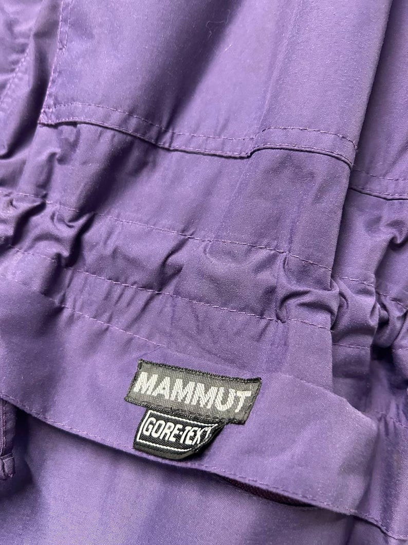 May include: Close-up of a purple garment with stitched seams and a drawstring. A black and white label reads "MAMMUT GORE-TEX". The fabric appears to be a durable, weather-resistant material, likely for outdoor wear.