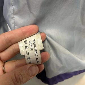 May include: Close-up of a white clothing tag with black text, held against a light blue fabric. The tag lists fabric composition: 70% POLYAMID 6.6 and 30% PTFE. Care symbols are also visible.