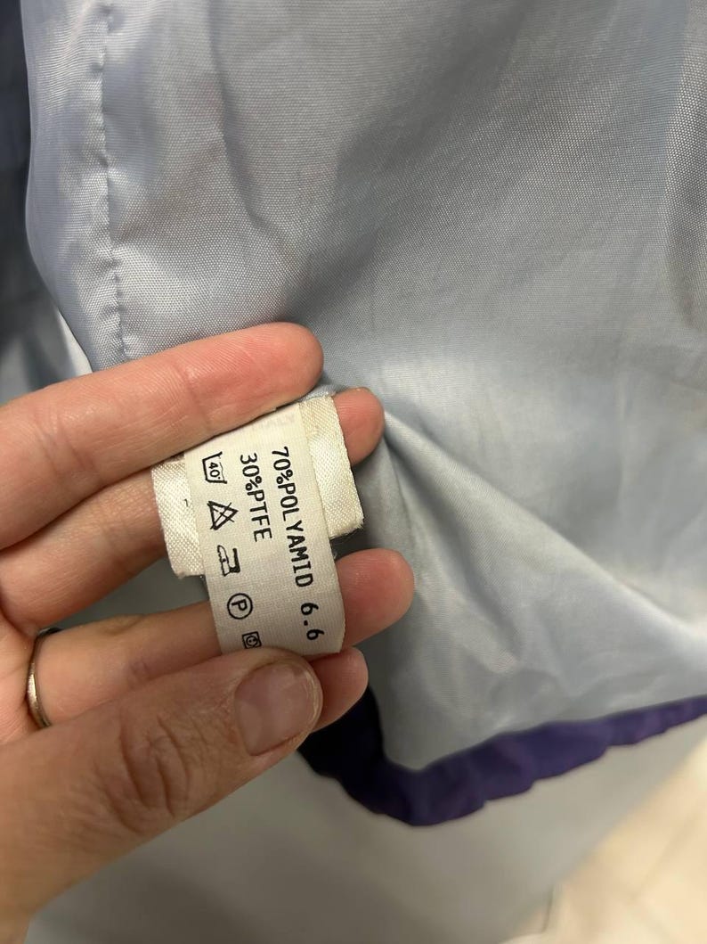 May include: Close-up of a white clothing tag with black text, held against a light blue fabric. The tag lists fabric composition: 70% POLYAMID 6.6 and 30% PTFE. Care symbols are also visible.
