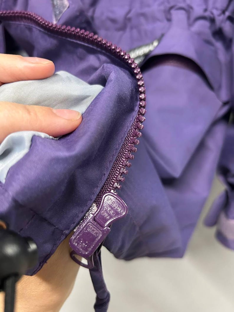 May include: Close-up of a purple jacket with a zipper. The zipper is a darker shade of purple and has the brand name "Vision" printed on it. The jacket's interior lining is a light gray color.