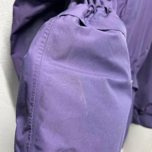 May include: Close-up of a purple jacket sleeve. The sleeve is gathered at the cuff and has a smooth, slightly shiny texture. The stitching is visible along the seams. The jacket appears to be made of a lightweight, water-resistant material.